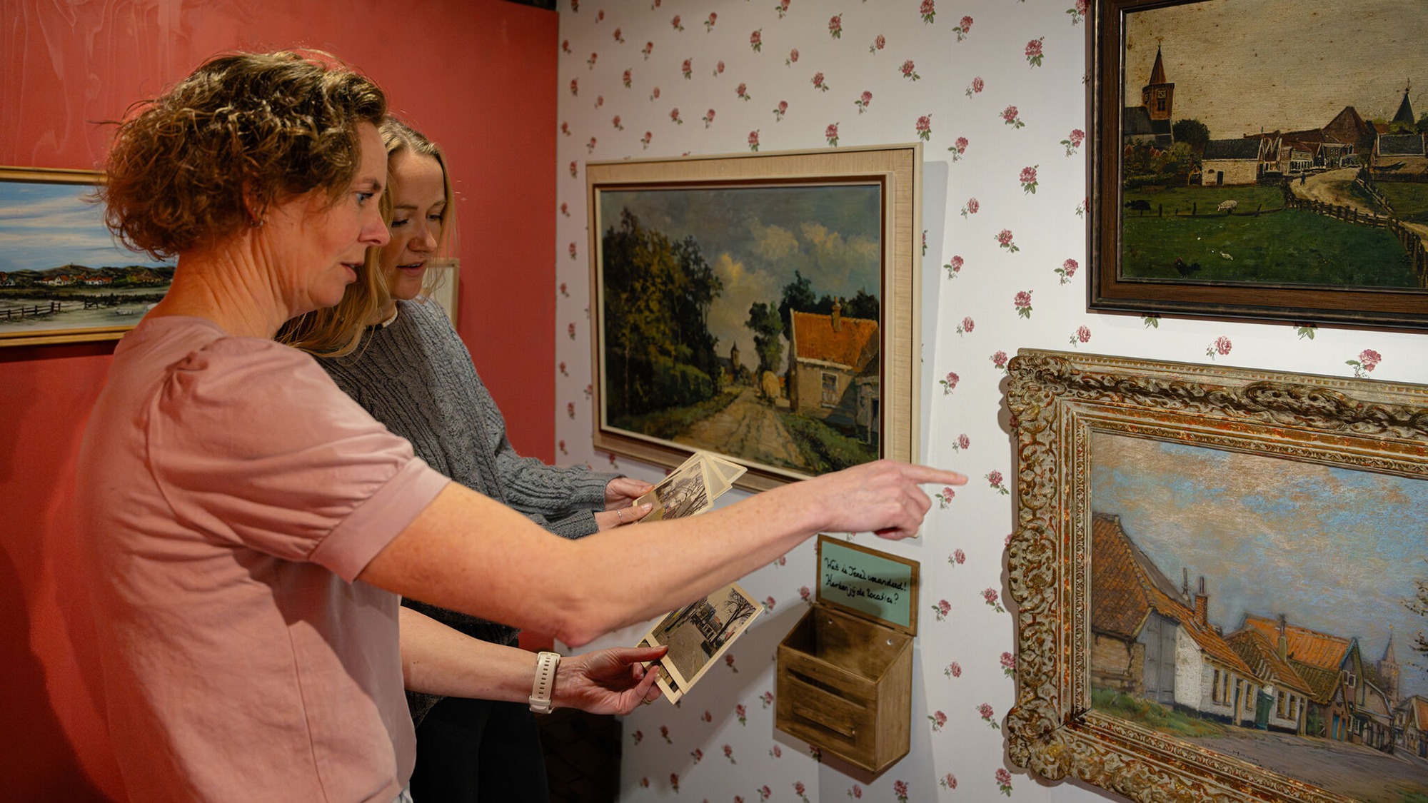 Twee dames vergelijken oude ansichtkaarten met schilderijen.