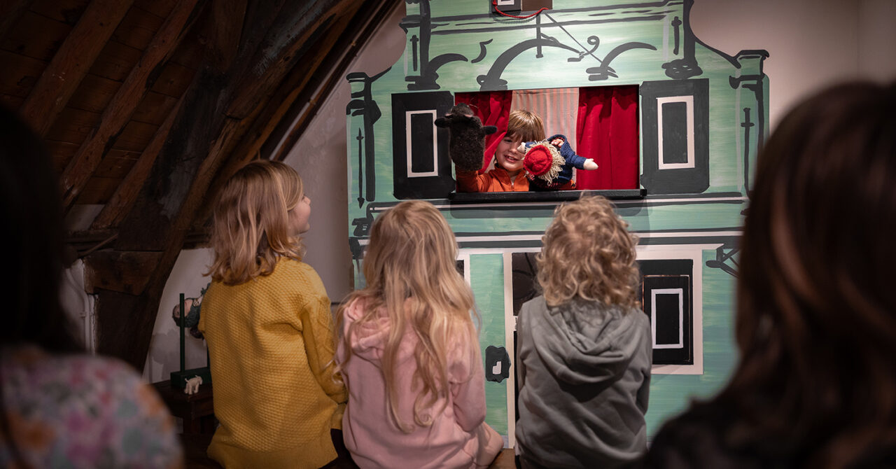 Kinderen kijken naar een jongetje dat met de poppen in de poppenkast speelt.
