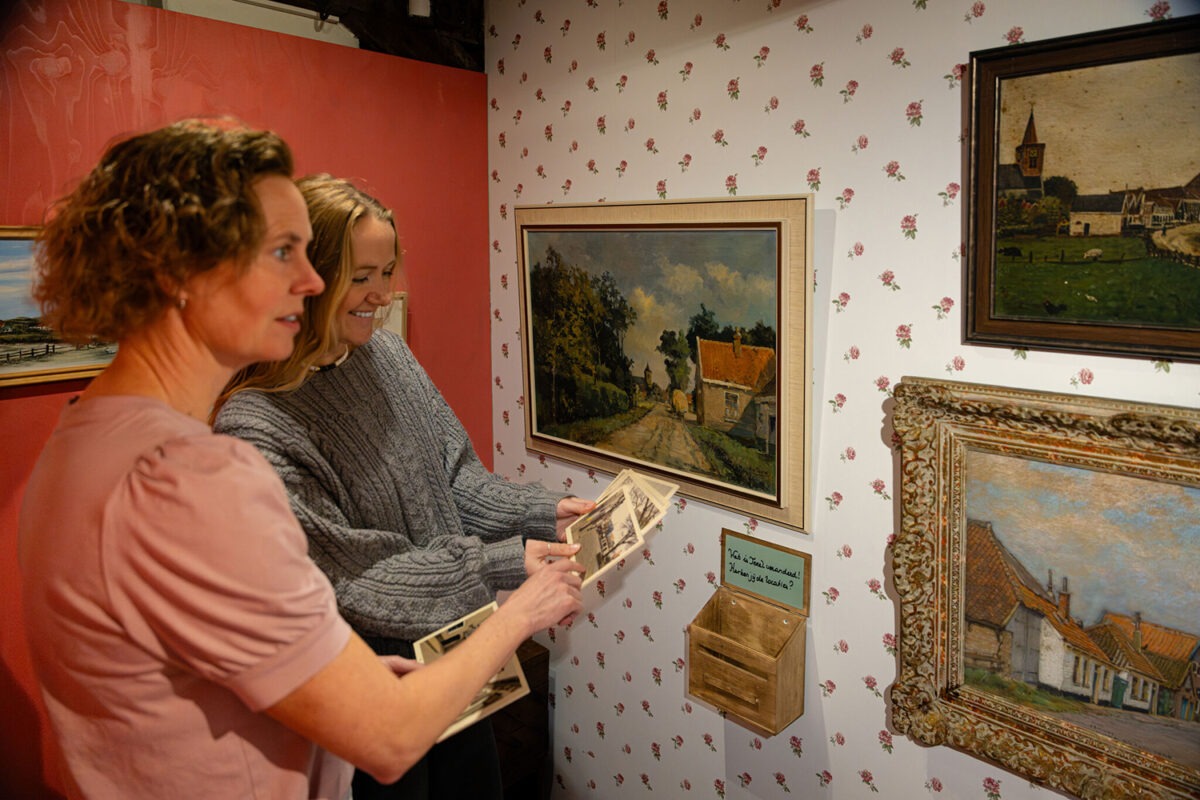 Twee dames vergelijken oude ansichtkaarten met schilderijen aan de muur.