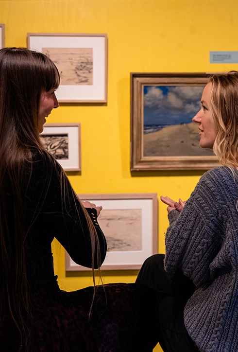 Twee jonge vrouwen, vanaf de rug gezien. Ze praten met schilderijen op de achtergrond.