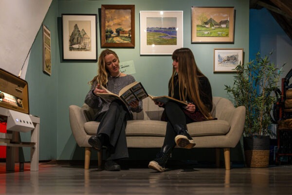 Twee jonge vrouwen kijken in een fotoboek, zittend op een bank met schilderijen erboven