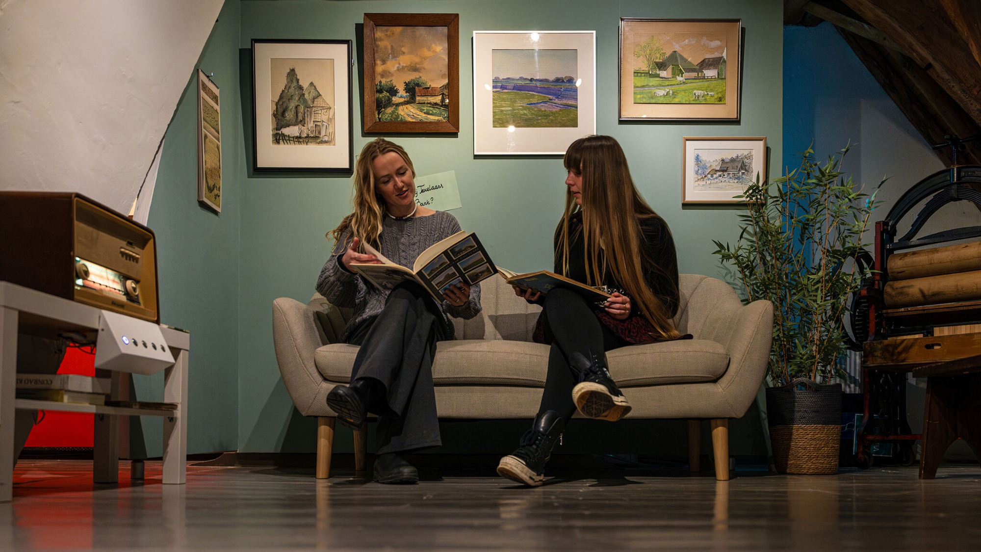 Twee jonge vrouwen kijken in een fotoboek, zittend op een bank met schilderijen erboven