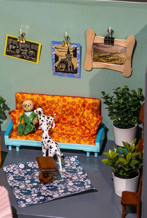 Een hand houdt een lapje vast voor een ingerichte poppenhuiskamer.